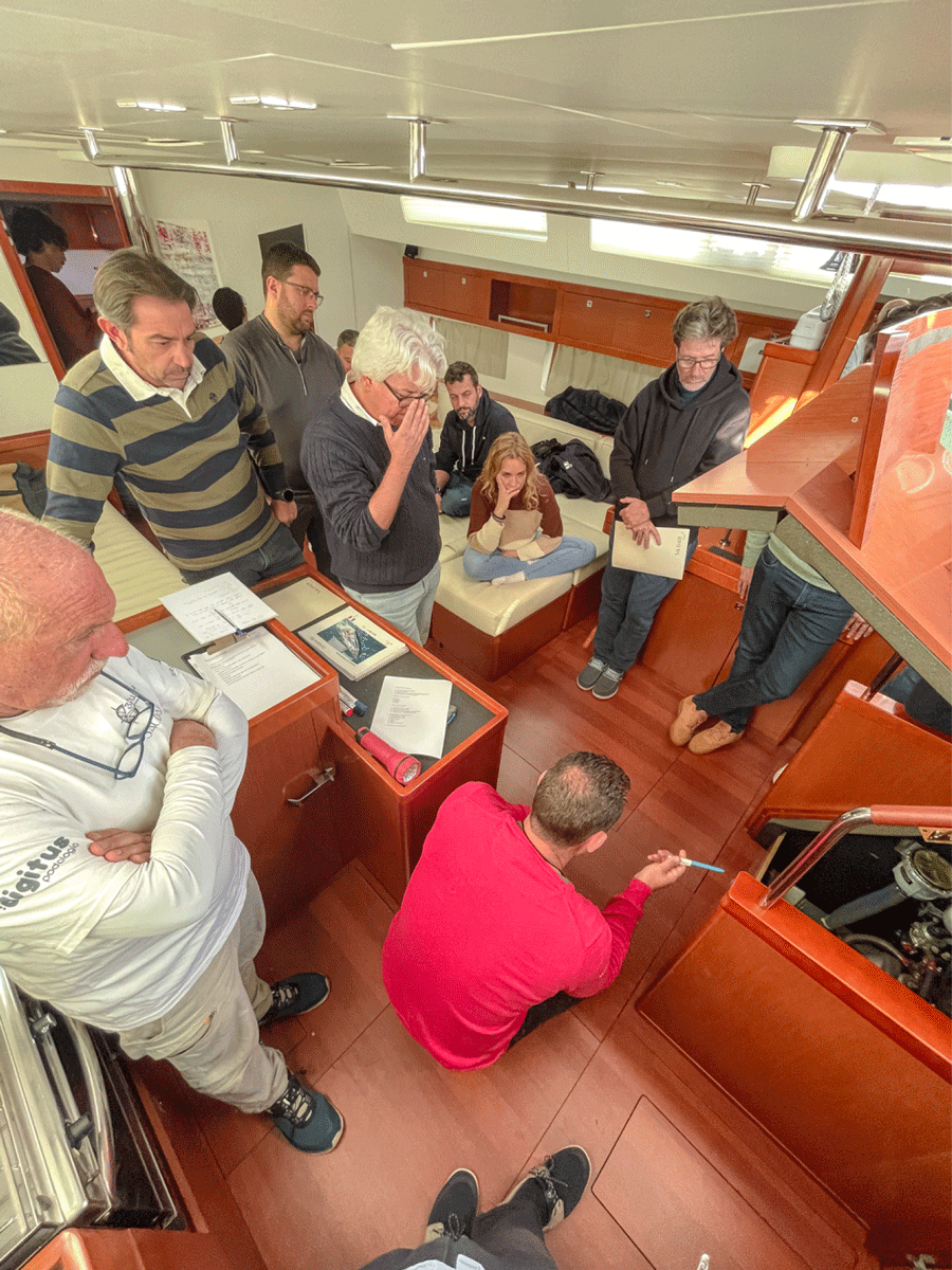 Un grupo de personas están reunidas dentro de la cabina de un barco, algunas sentadas y otras de pie. Una persona con una camiseta roja señala algo fuera de cuadro, probablemente durante el Curso de Navegación: Mecánica. Una computadora portátil y papeles son visibles sobre la mesa, resaltando el ambiente enfocado de formación a bordo.