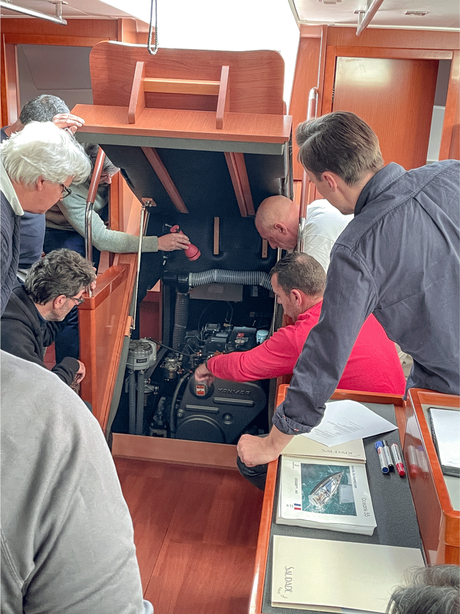 Varias personas se reúnen y trabajan en el compartimento abierto del motor de un barco dentro del interior de un barco de madera como parte del Curso de Navegación: Mecánica. Algunas personas examinan el motor de cerca mientras otras observan, beneficiándose de la formación a bordo en La Marina de València.