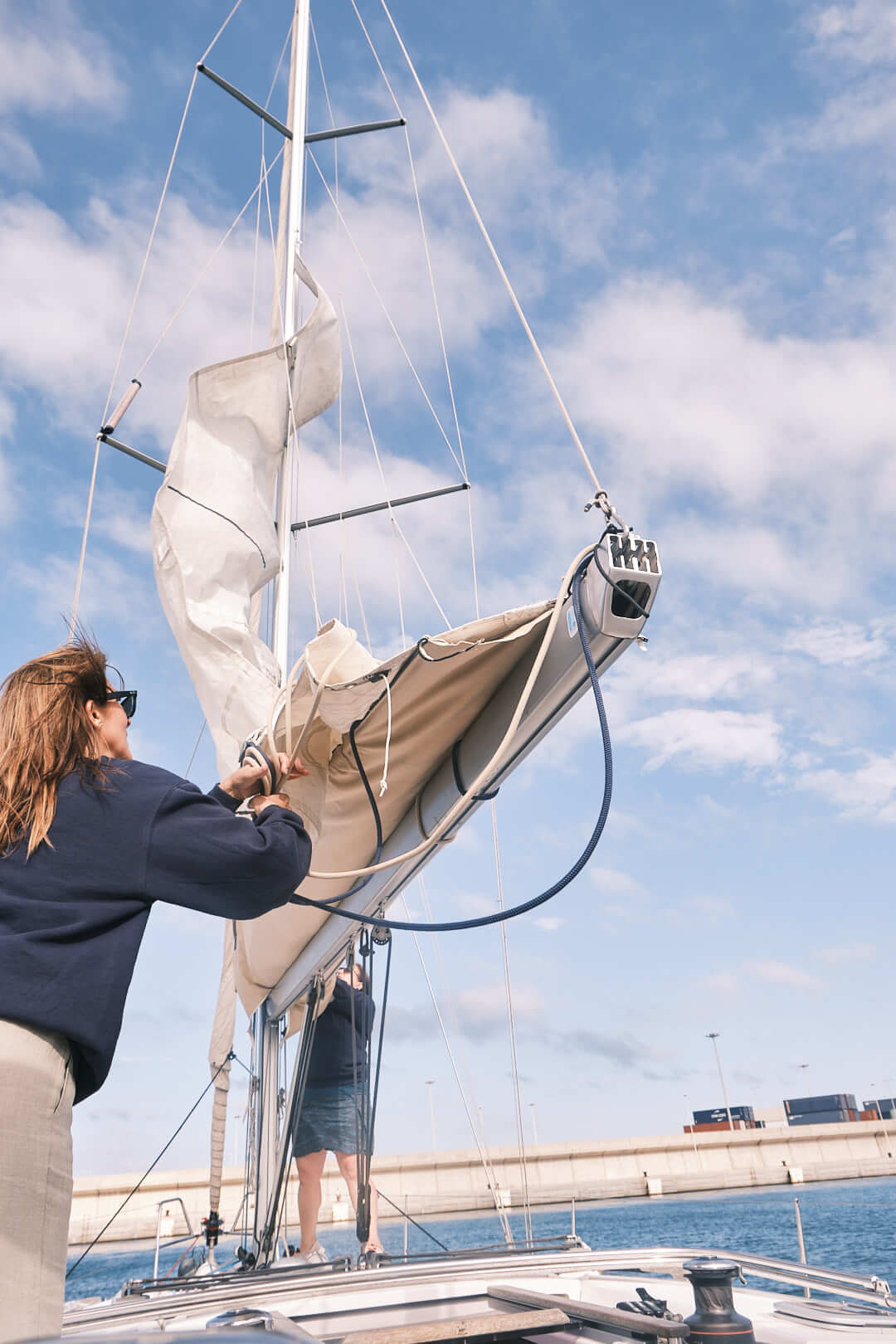 Una persona asegura la vela en un velero atracado bajo un cielo parcialmente nublado en La Marina de València, participando en el Curso de Navegación: Aparejado y Trimado de Velas.