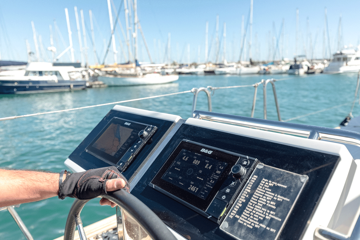 Una persona navega en un barco mientras ve datos de dos sistemas de navegación, con La Marina de València y varios yates y veleros al fondo, lo que ilustra el aprendizaje práctico del Curso de Navegación: Cartas náuticas y plotter combinado con formación en clase y a bordo.