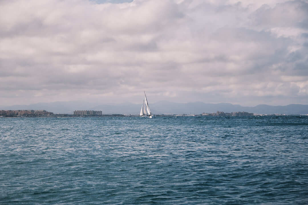 Un velero se desliza sobre una vasta masa de agua bajo un cielo nublado, con edificios distantes y montañas visibles en el horizonte. Quizás forme parte del Curso de Navegación: Meteorología que organiza La Marina de València.