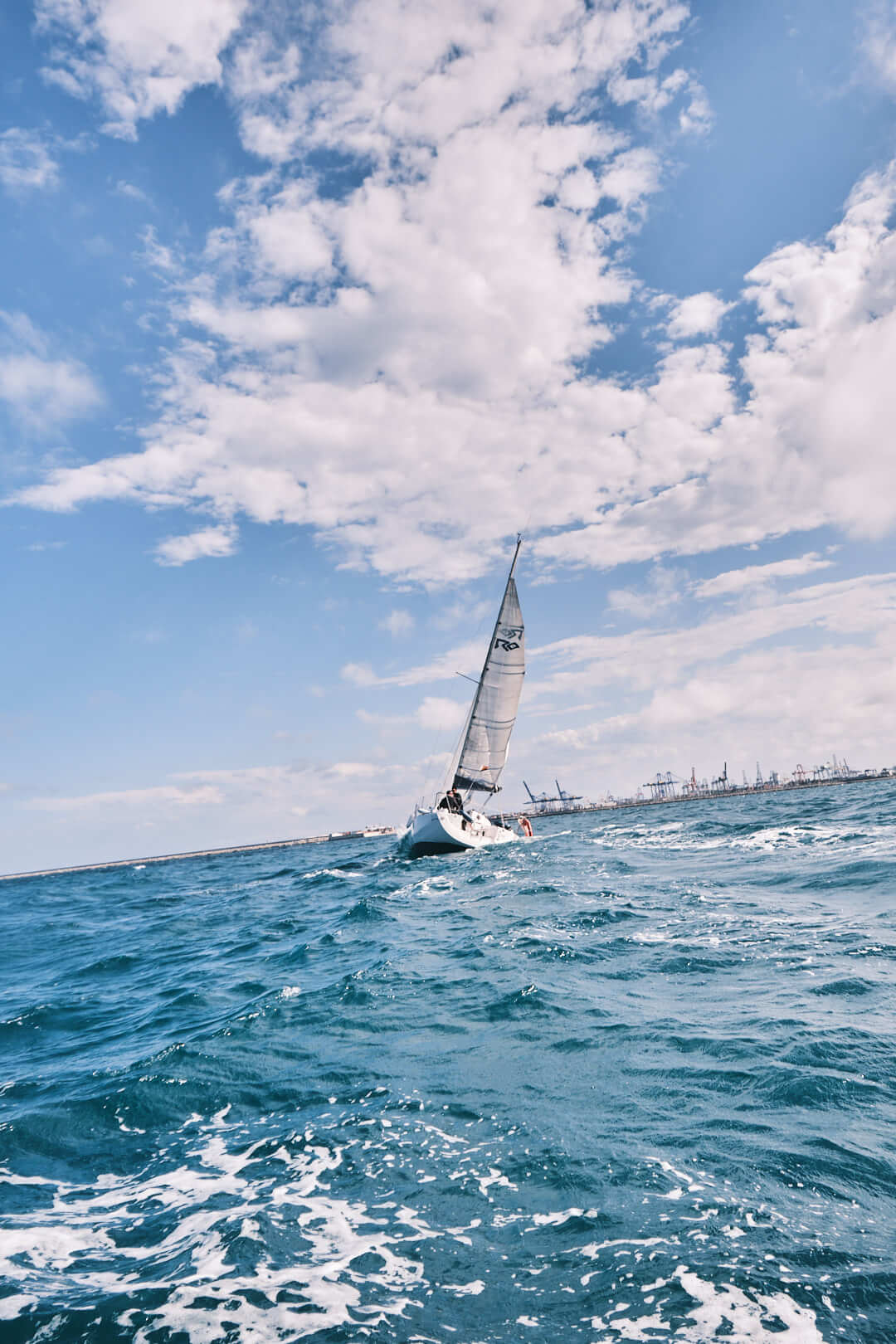 Un velero con la vela erguida se desliza sobre un mar azul bajo un cielo parcialmente nublado, con el telón de fondo del litoral industrial de La Marina de València, tal y como muestra el "Curso de Navegación: Meteorología.