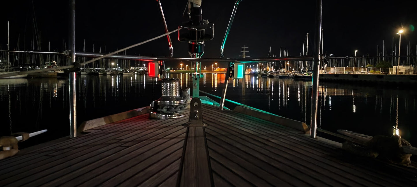 Vista nocturna desde la proa de un barco atracado en La Marina de València, con luces rojas y verdes iluminando el entorno y reflejos de otros barcos visibles en el agua, encapsulando perfectamente el ambiente sereno que a menudo destaca Oscar García Flores durante su *Curso de Navegación : Sesiones nocturnas*.