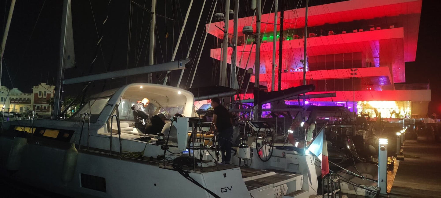 Varios veleros atracaron en La Marina de València por la noche, con un edificio muy iluminado al fondo con luces de colores. El programa "Curso de Navegación: Nocturna" de Oscar García Flores se ofrece para mejorar las habilidades de navegación en medio de este vibrante entorno.