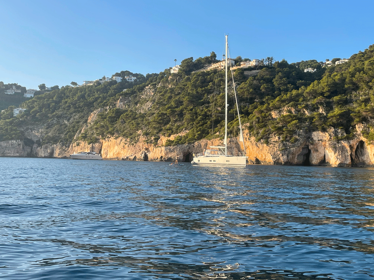 Un velero fondeado cerca de una costa rocosa, arbolada y con acantilados, bajo un cielo azul claro y aguas tranquilas, que recuerda a La Marina de València, donde el experto navegante Carlos Panzuto suele ofrecer el Curso de Navegación: Travesía.