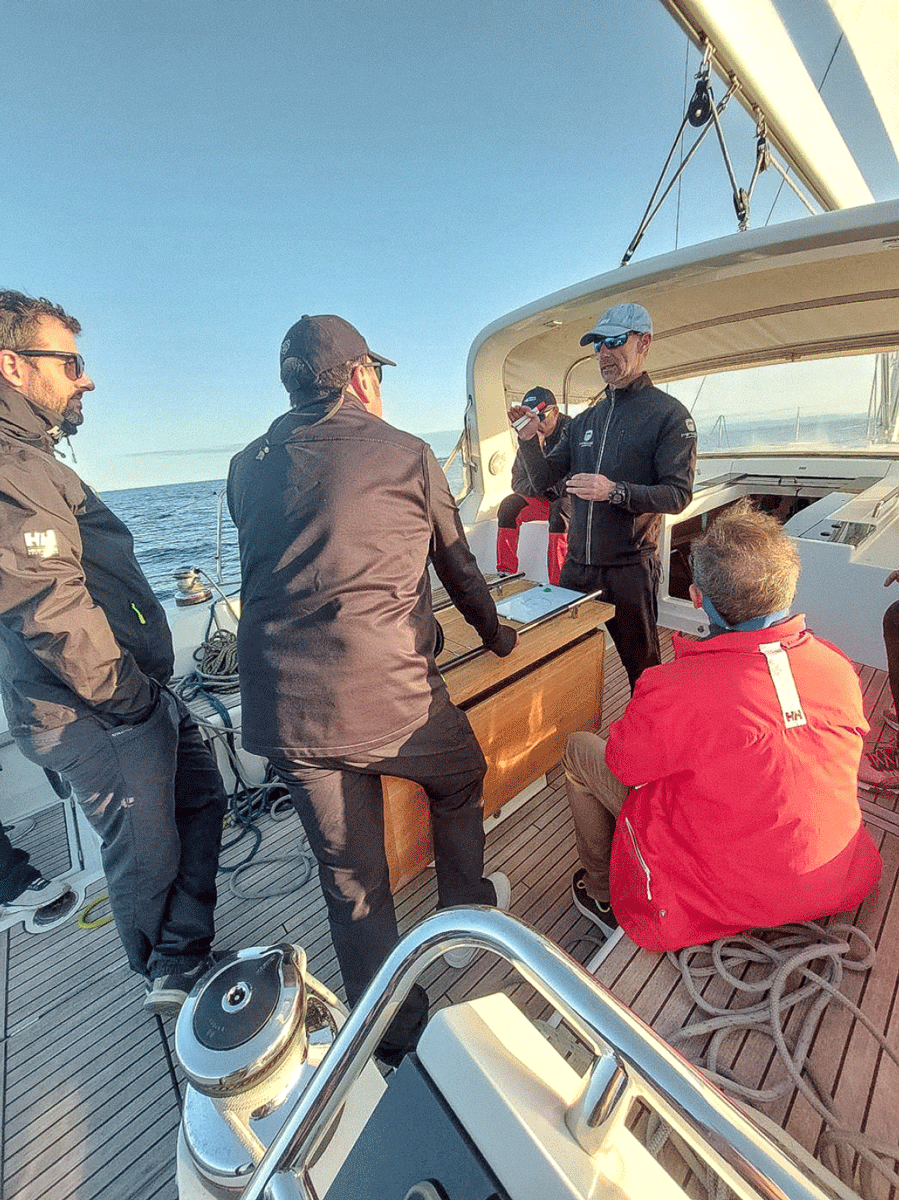 Un grupo de personas con equipo de navegación están reunidas en un yate en el océano cerca de Marina de València. Una persona se pone de pie y habla mientras otros escuchan atentamente, como parte de su Curso de Navegación: Velas Portantes. El escenario es tranquilo con cielos despejados.